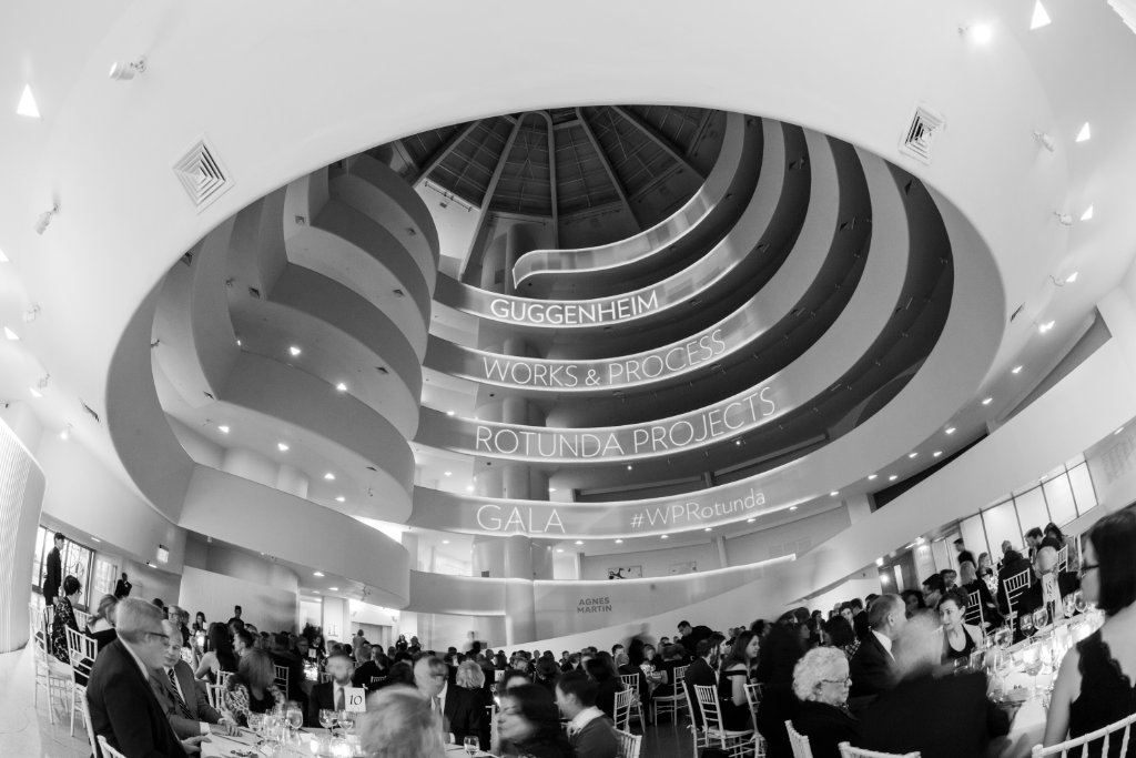 Stadium-scale corporate tech event with dramatic stage lighting, silhouetted presenter, and audience of glowing smartphones