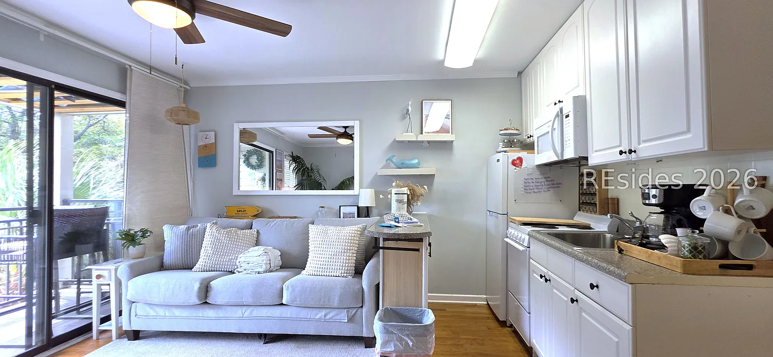 Open-concept living and kitchen of Pebble Beach Lodge with light gray sofa, white cabinetry, and sliding glass door to a balcony