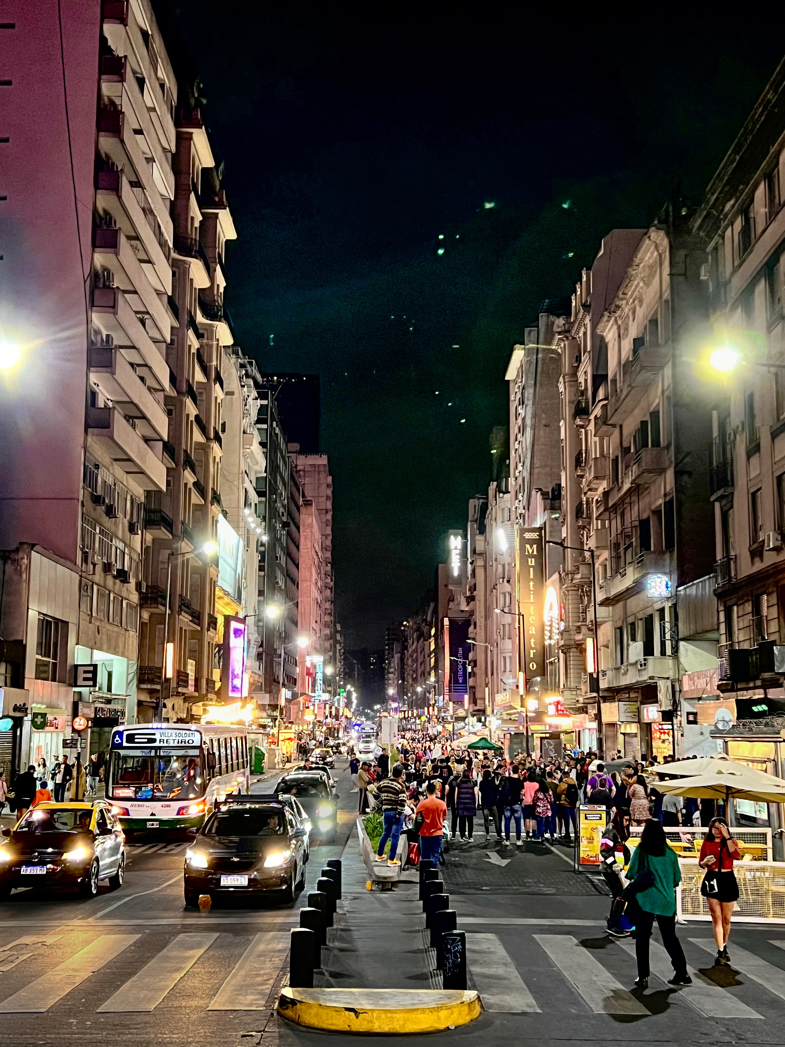 A vibrant night scene on Corrientes Avenue in Buenos Aires, with bustling traffic, illuminated theater signs, and crowds on the sidewalk