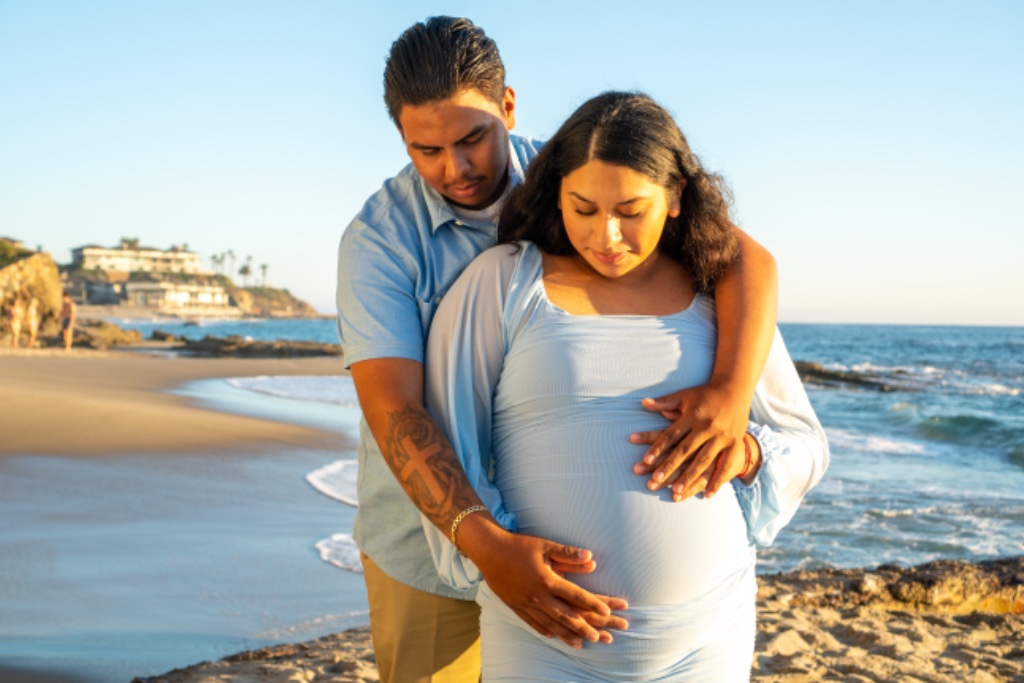 Couple embracing on beach, man's hands on pregnant belly, Laguna Beach cliffs behind