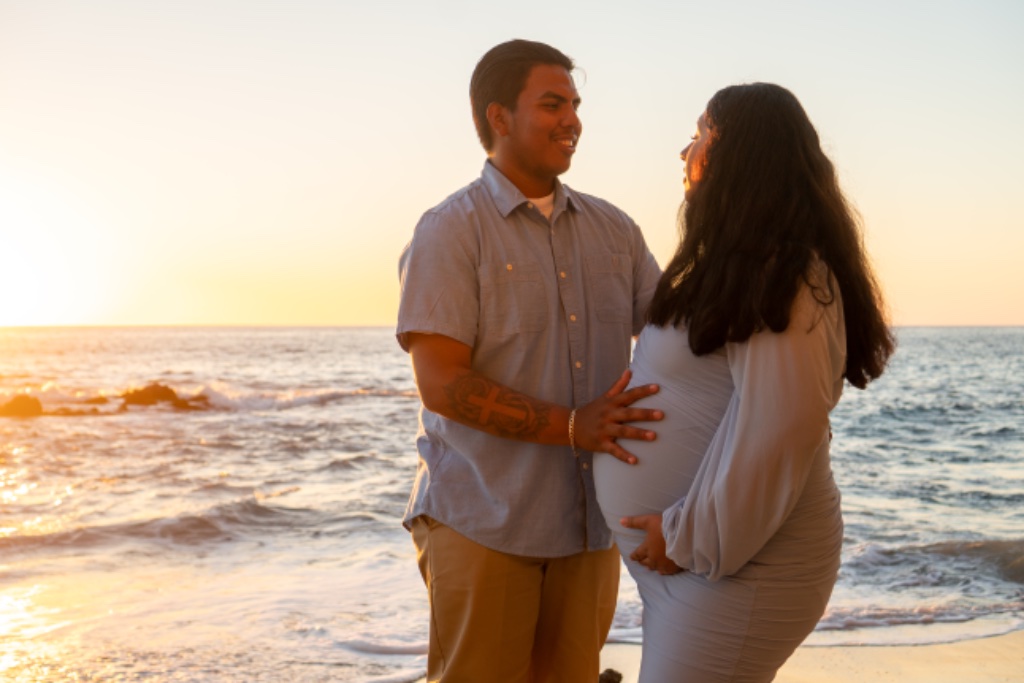 Couple in profile on beach at golden hour, hands on baby bump