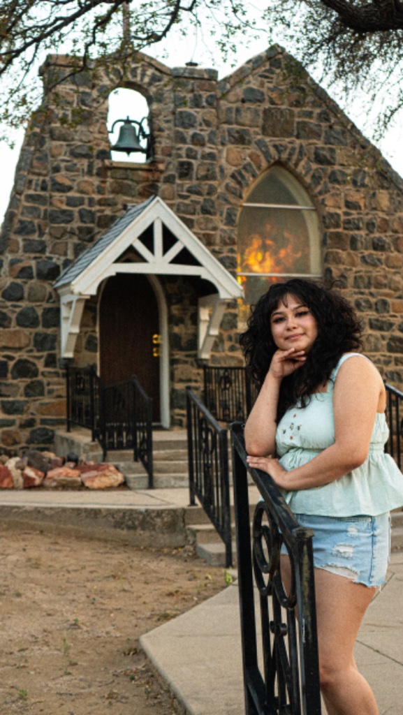 Graduate in mint green top leaning on black iron railing with stone chapel and bell tower