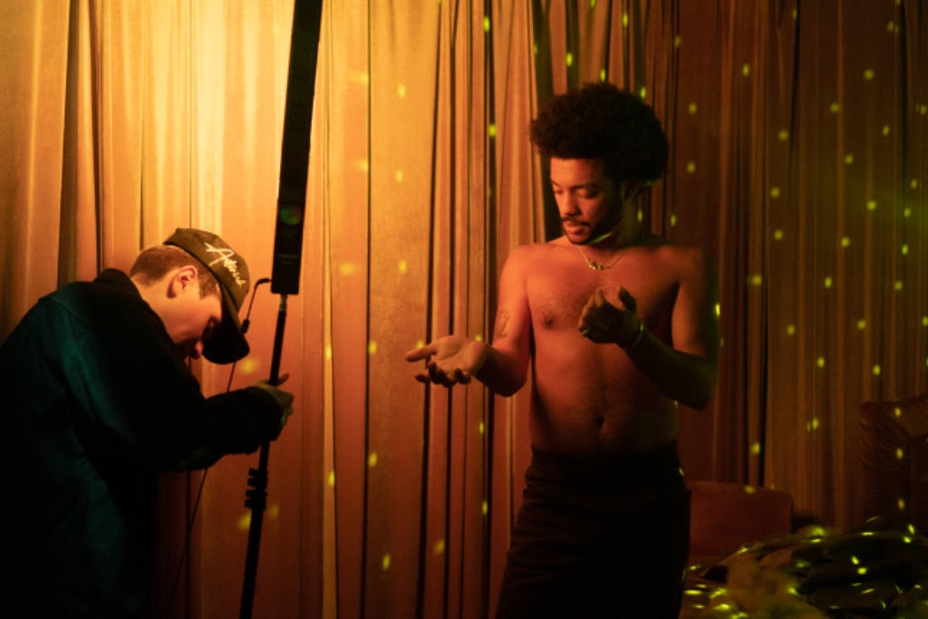 Felix Ames shirtless with photographer holding light wand, golden lighting