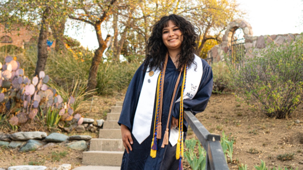 Graduate leaning on black metal handrail with stone church and bell tower in background