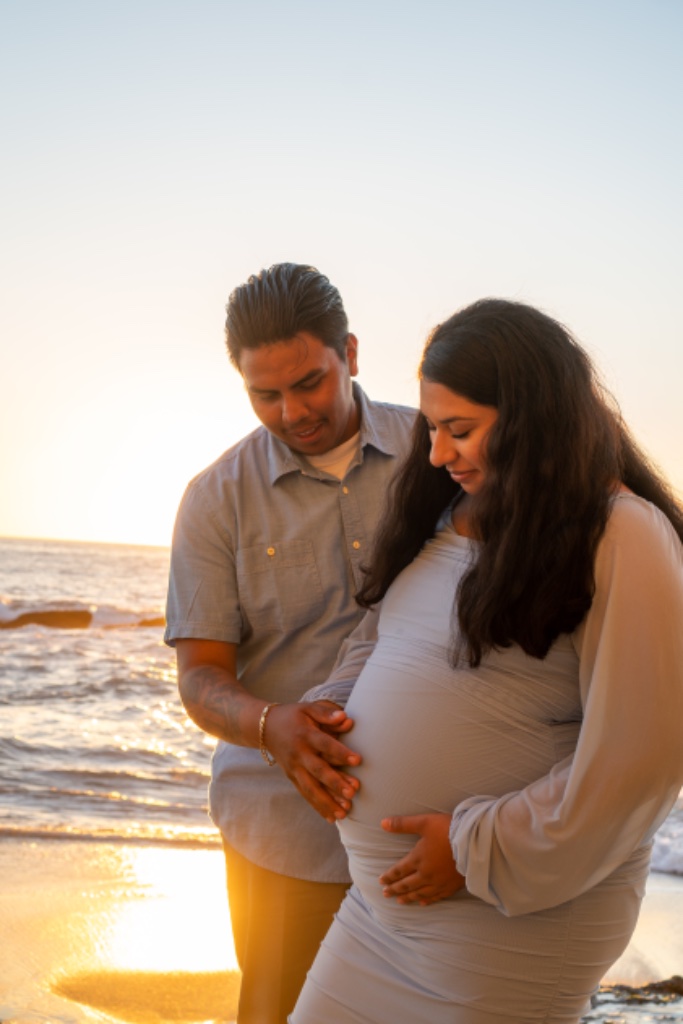 Couple looking at baby bump together on beach at warm sunset