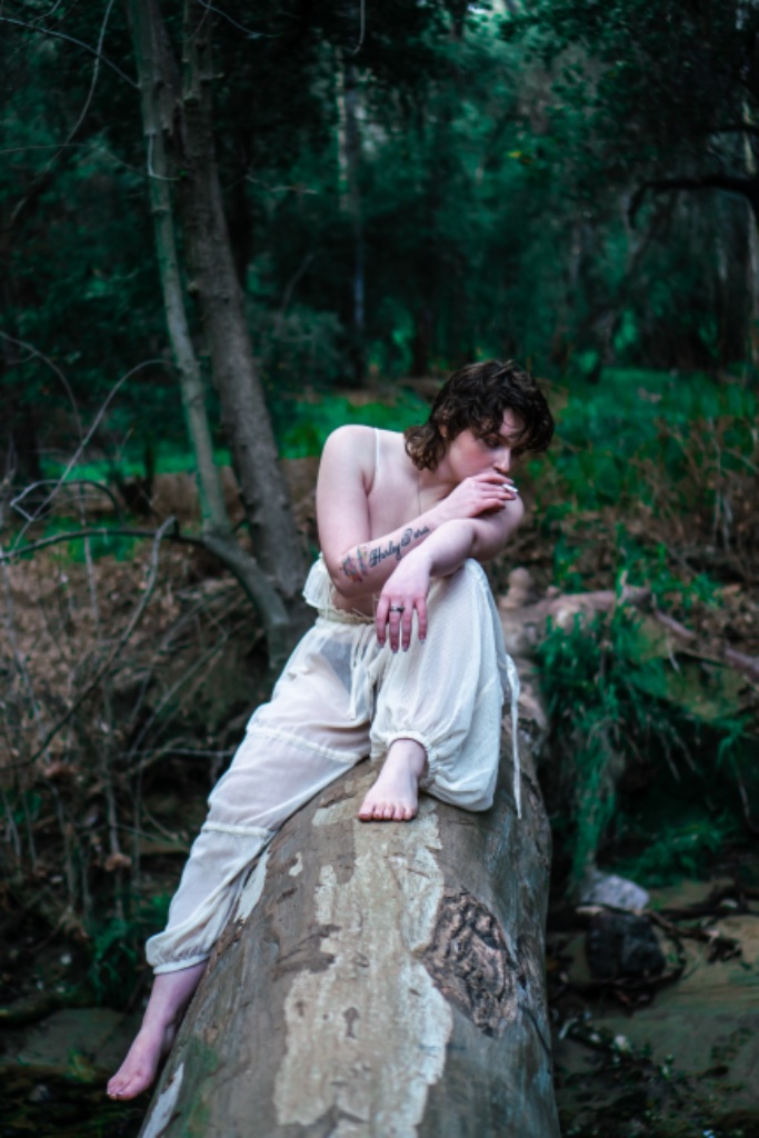 Woman sitting on tree log, cigarette to mouth, Harley Paris tattoo visible