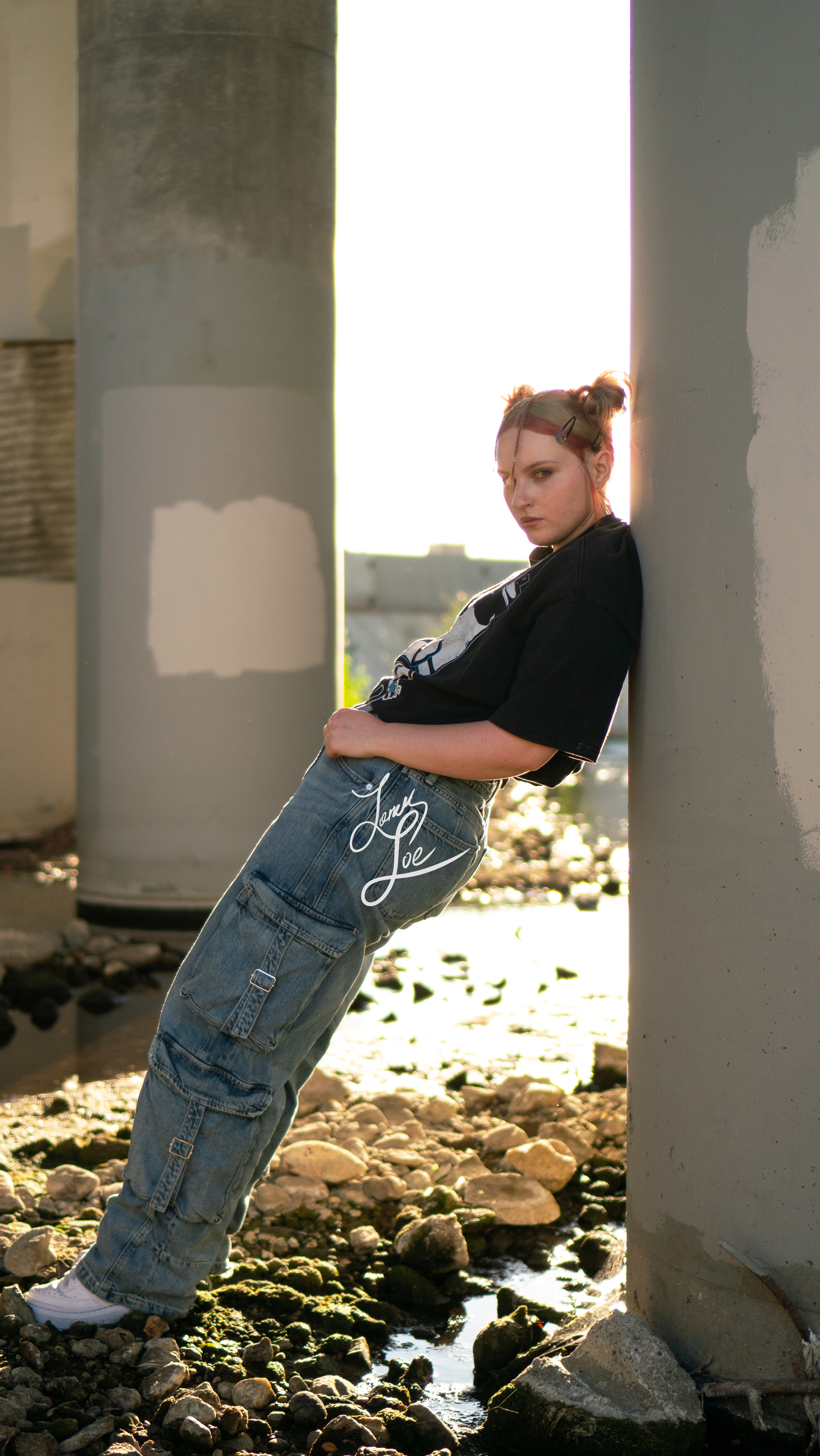 Woman leaning between two concrete pillars, cargo jeans and graphic tee, backlit