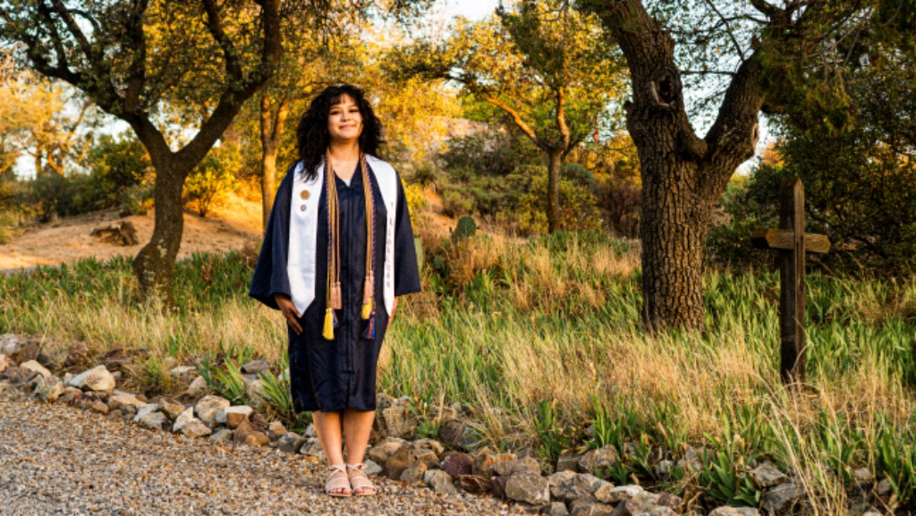 Graduate in navy gown walking on gravel path with wooden cross in background at golden hour