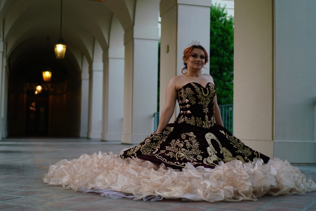 Quinceañera seated gracefully in a dramatic ball gown under archway lights at dusk