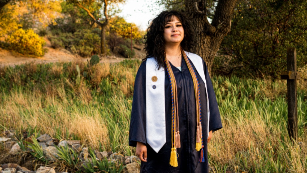 Full-length portrait of graduate in navy gown standing on gravel path with tree and cross