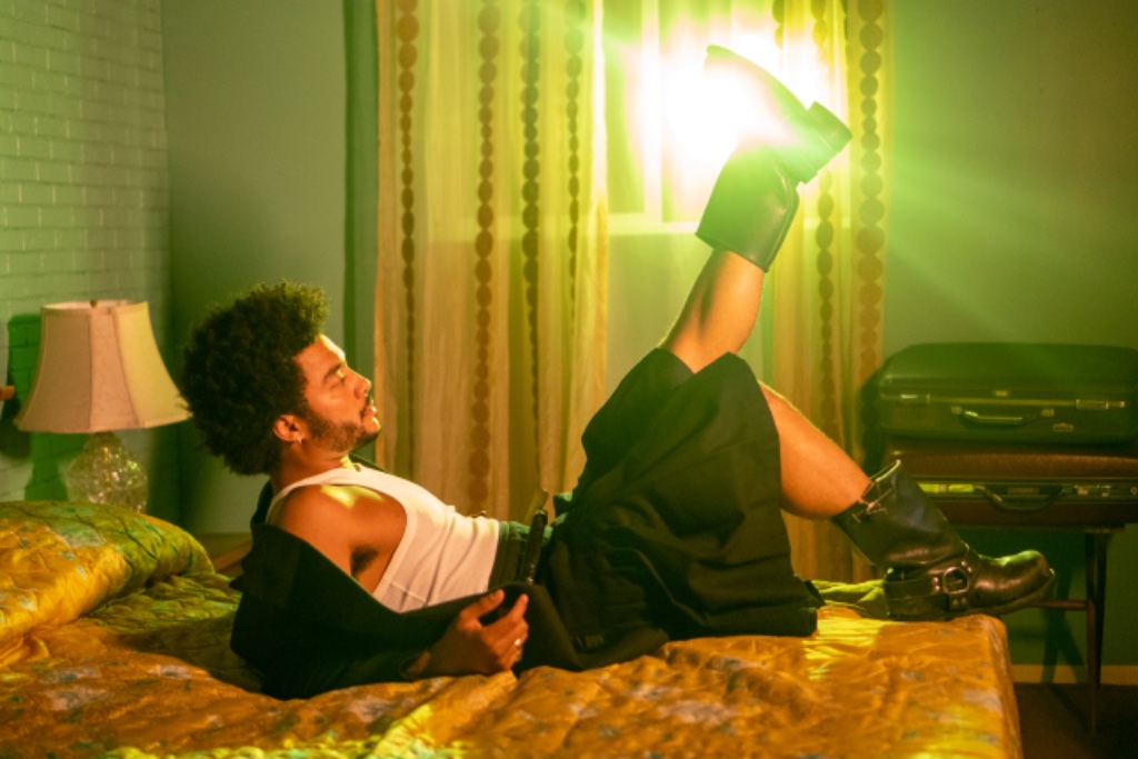 Felix Ames in white tank top and kilt on gold quilted bed with green lighting