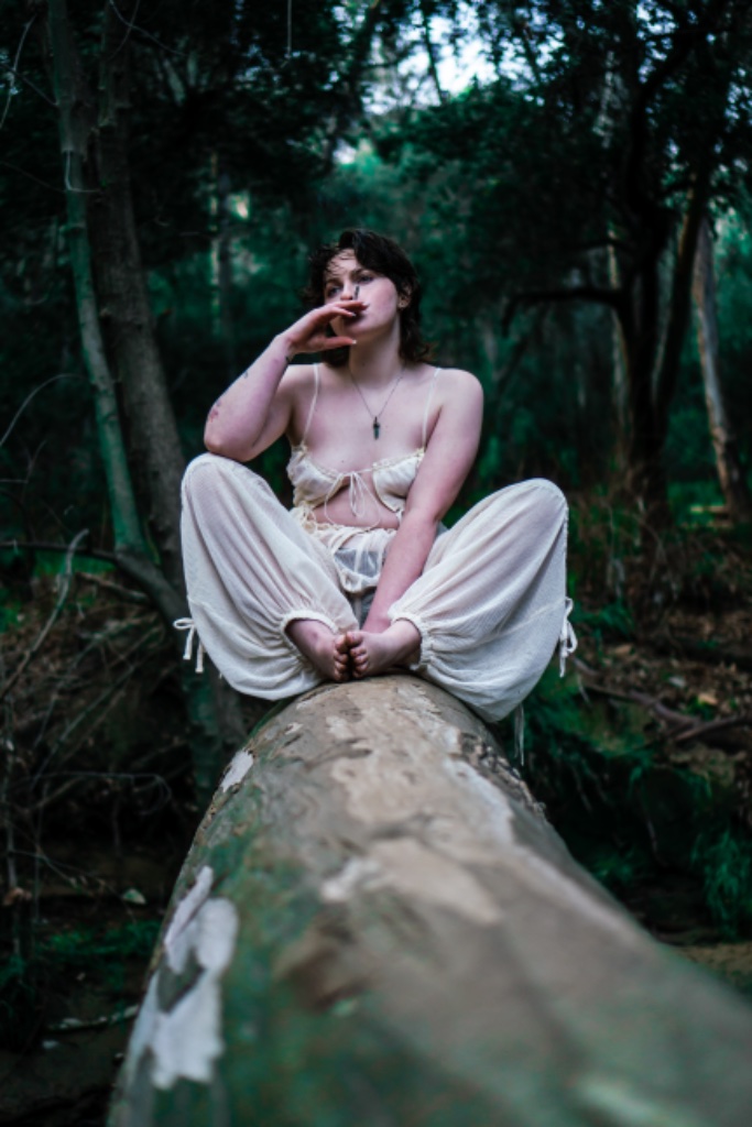 Woman sitting cross-legged on log, cigarette to mouth, looking away