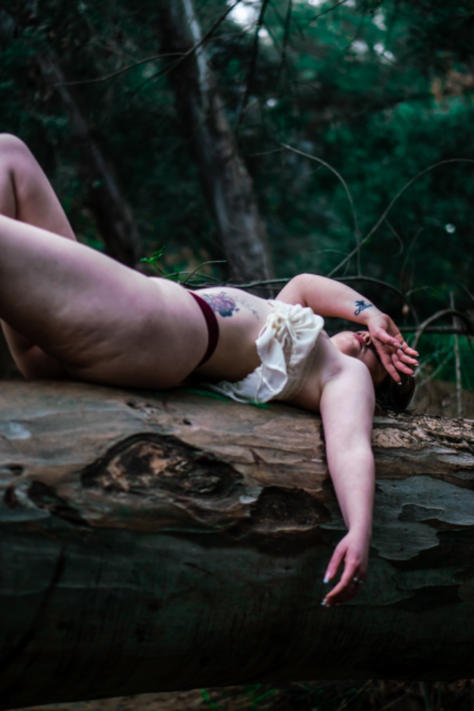 Woman lying on side on forest log, hand near face, flower tattoo on ribcage