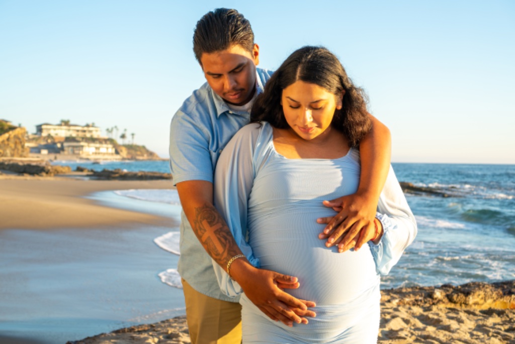 Couple on beach at golden hour, man embracing pregnant woman in blue dress