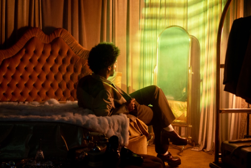 Felix Ames reclining on rust velvet bed in gold curtained room with mirror