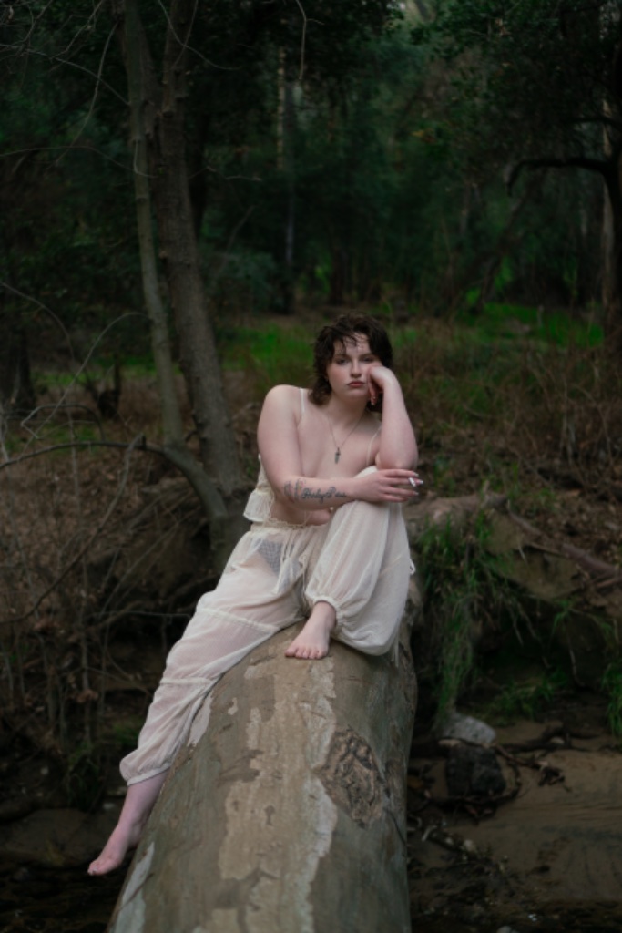 Person seated on fallen tree log in woods, cigarette in hand