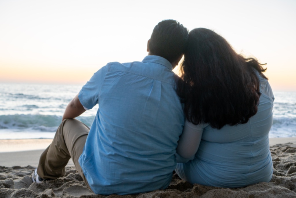 Couple sitting on beach at sunset, viewed from behind, golden sky