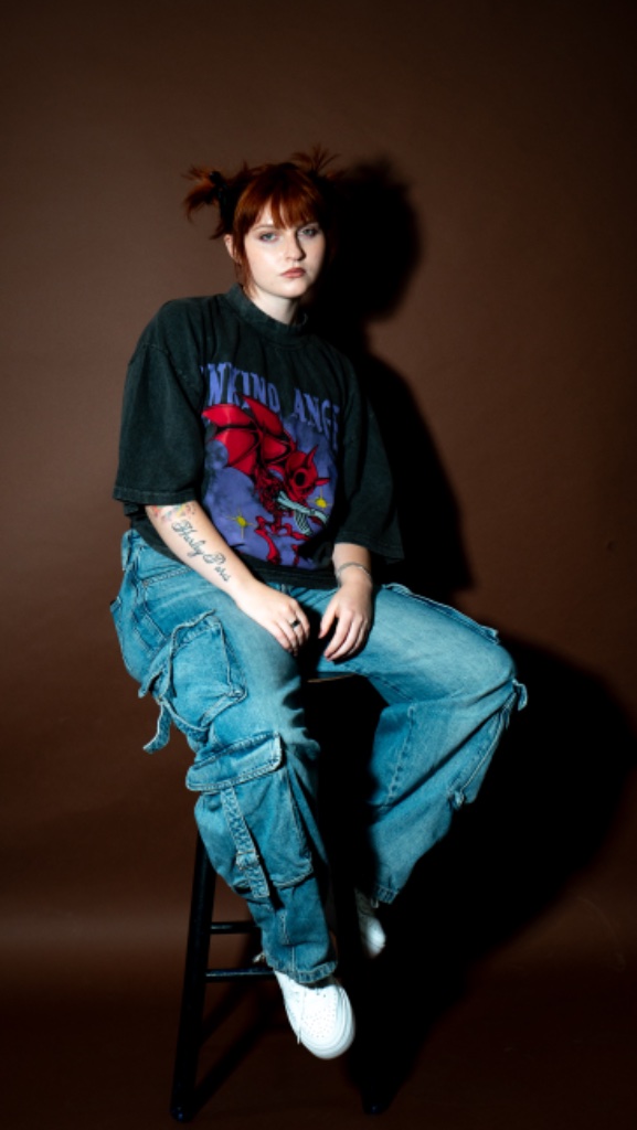 Female model seated on stool in graphic tee and cargo jeans, dark background