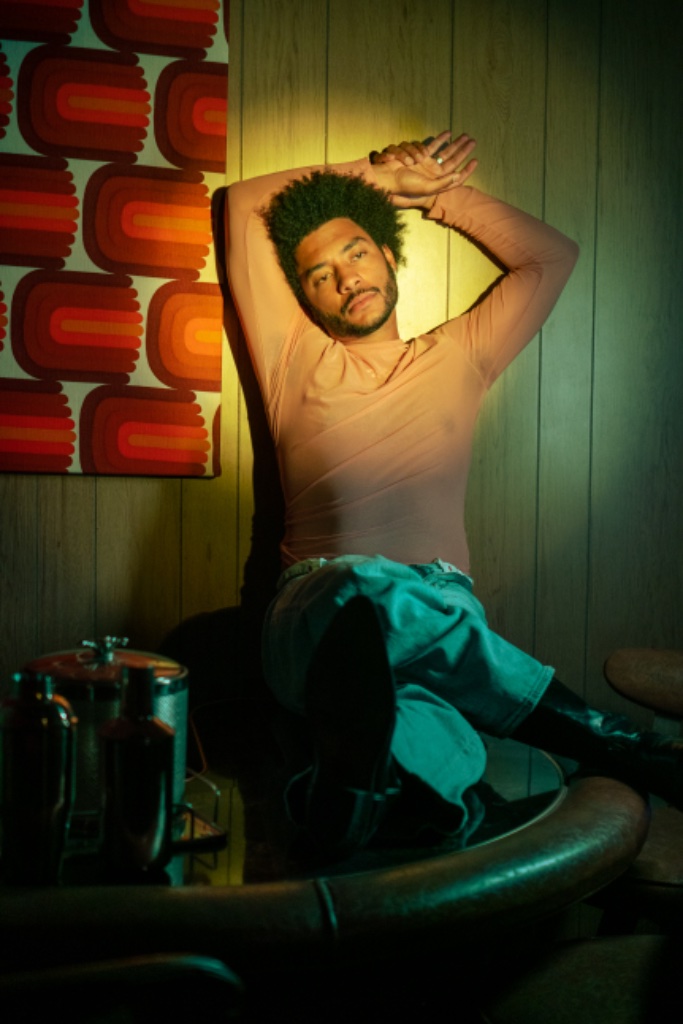 Felix Ames on glass table in pink top with arms raised, wood paneling