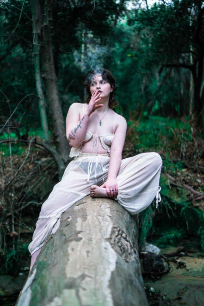 Woman sitting on fallen tree trunk, cigarette with smoke wisp, crystal pendant
