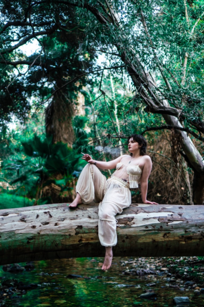 Woman sitting on log over stream, holding cigarette, full length portrait
