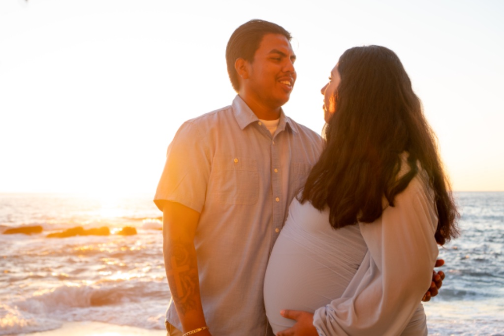 Couple at sunset with lens flare, man's cross tattoo visible, hands on baby bump