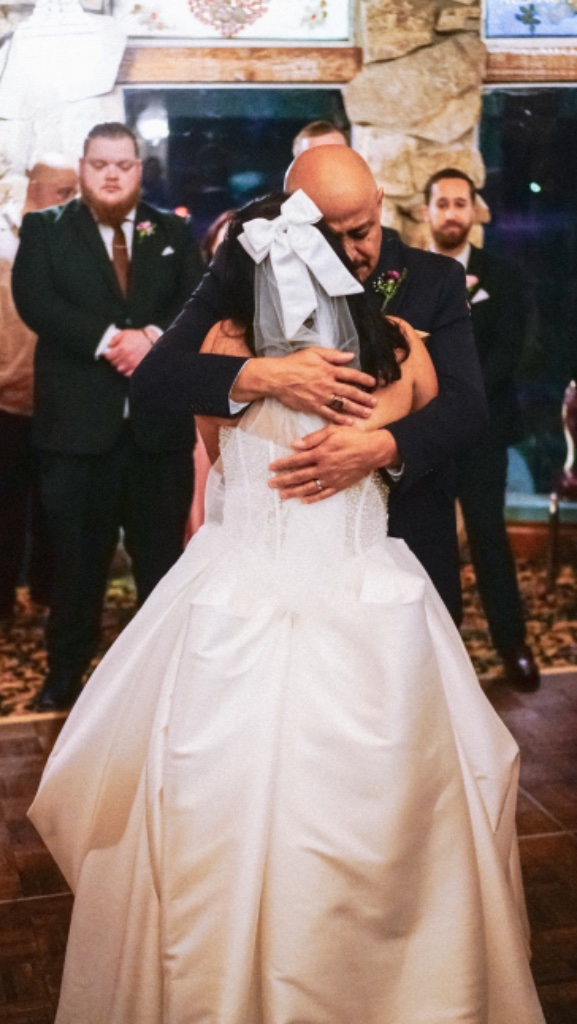 Bride with white bow veil hugging person in dark suit, groomsmen in background