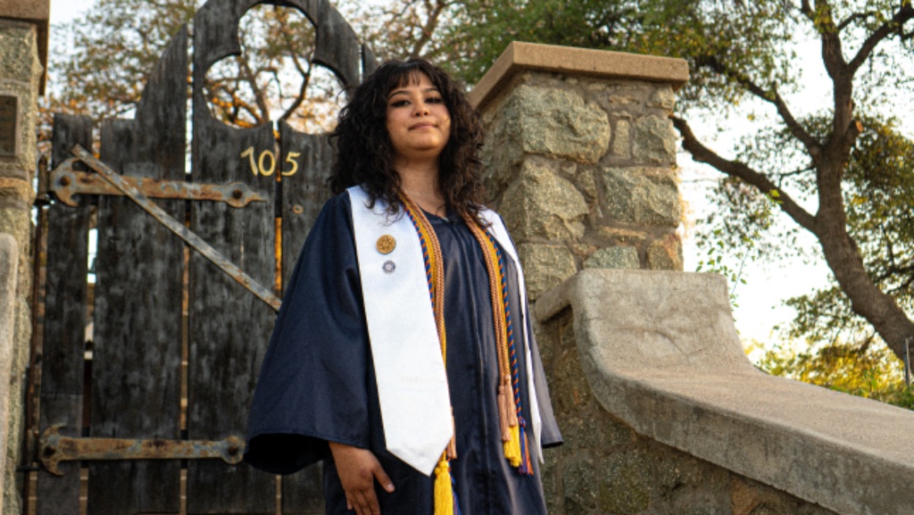 Graduate in navy gown and white stole standing before rustic wooden gate with Gothic arch