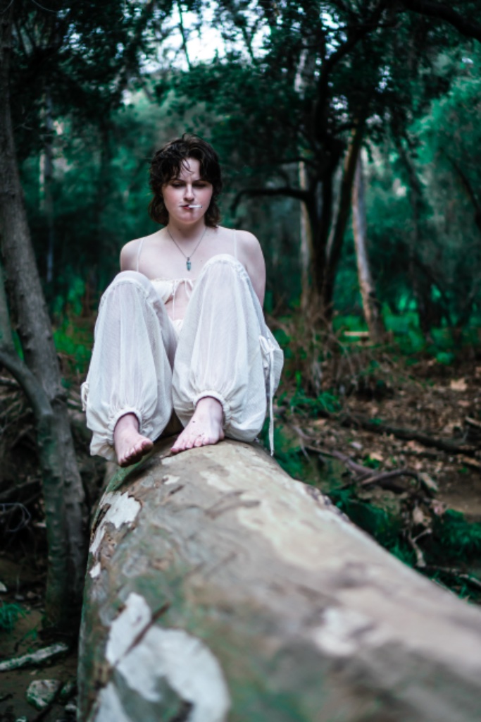 Woman sitting on log smoking cigarette, facing forward, barefoot
