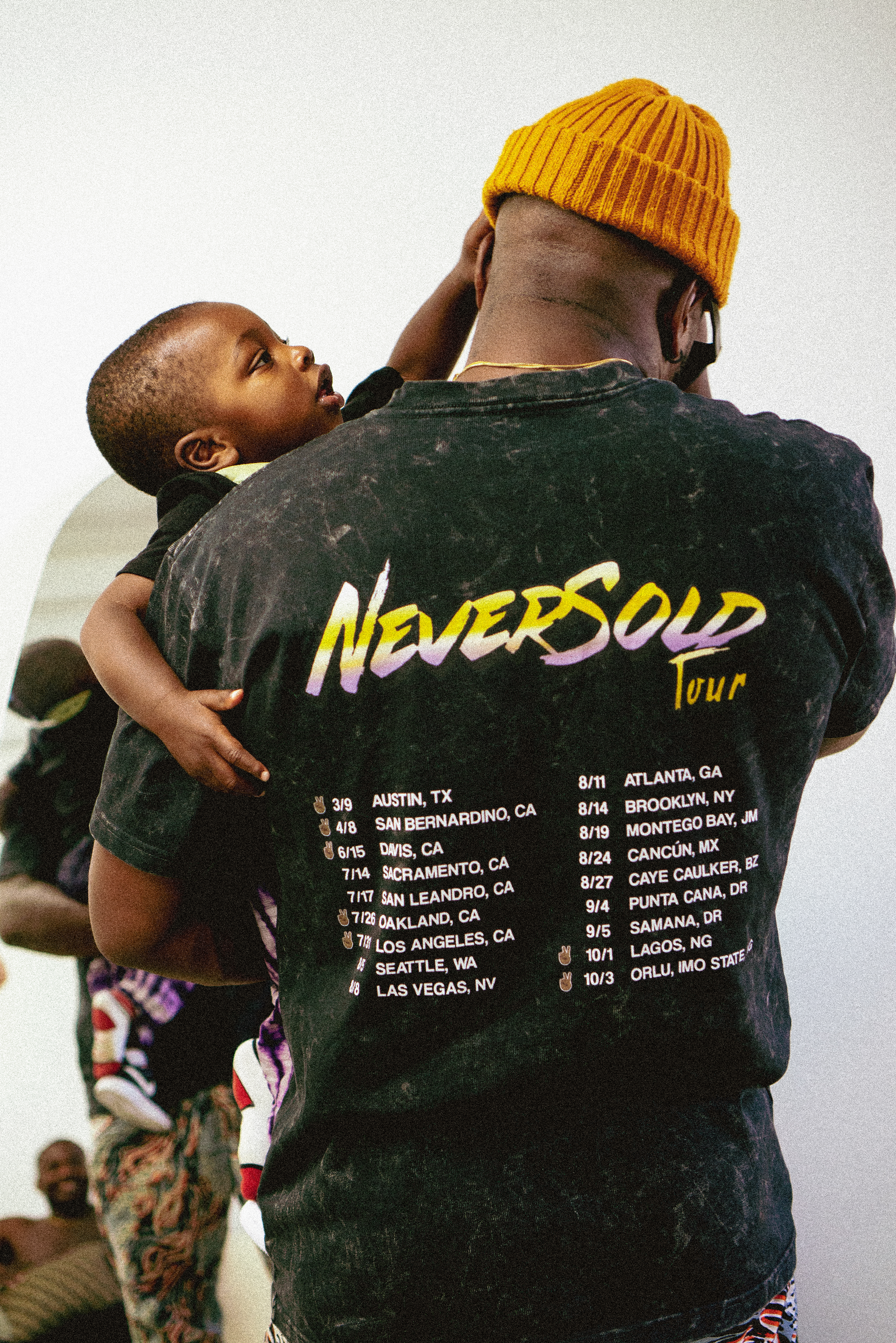 Man in NeverSold Tour tee holding child, yellow beanie, studio setting