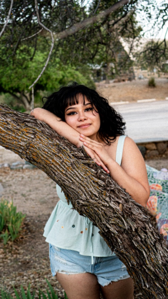 Graduate in mint green top leaning against large tree trunk in natural outdoor setting