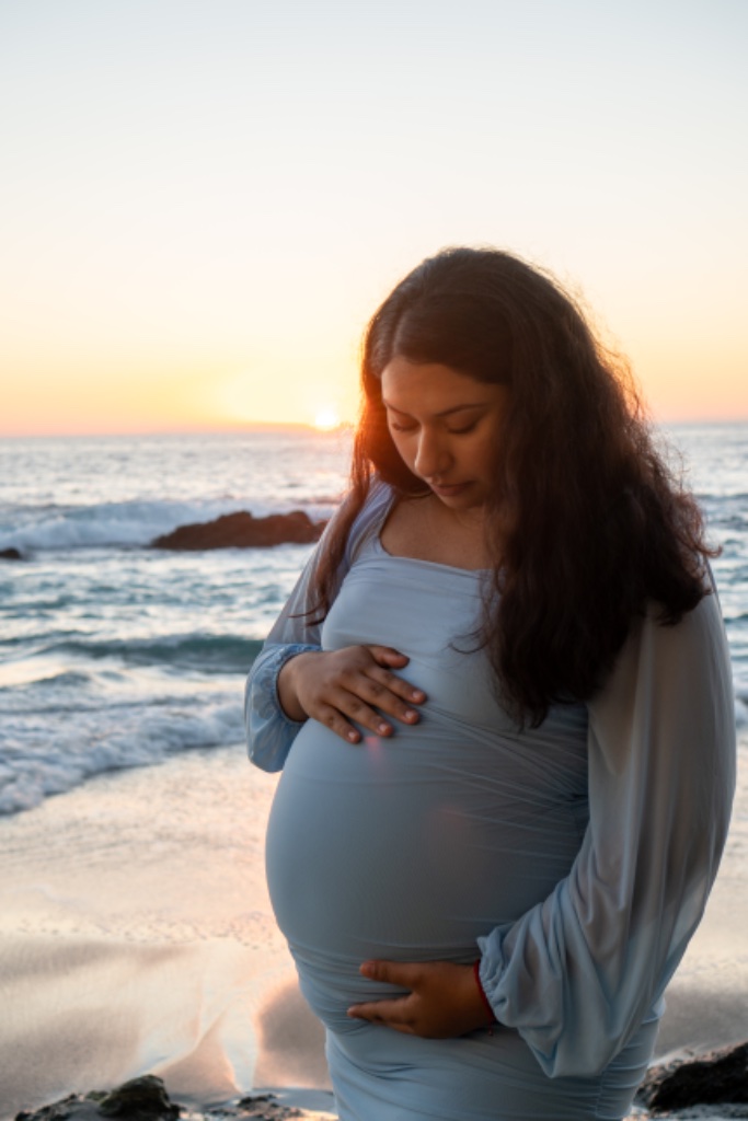 Pregnant woman in blue dress cradling belly on beach at sunset