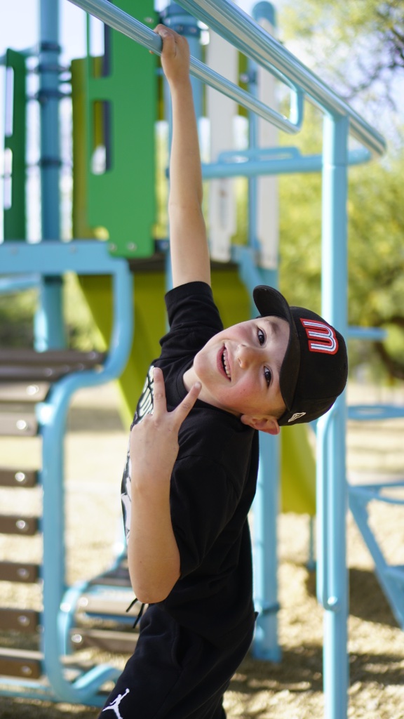 Boy hanging from playground bar smiling with cap, by Jose A. Palafox