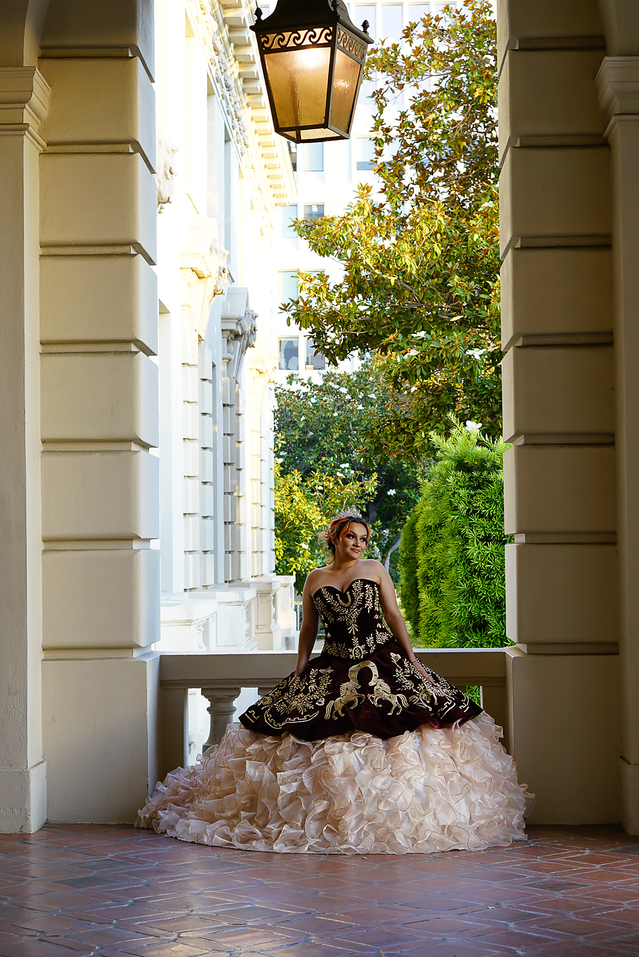 Quinceañera in ornate black and gold embroidered gown seated under historic archway with lantern