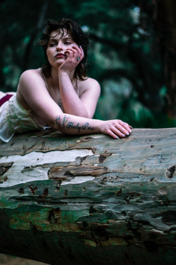 Woman lying prone on tree trunk, chin on hand, maroon undergarment visible