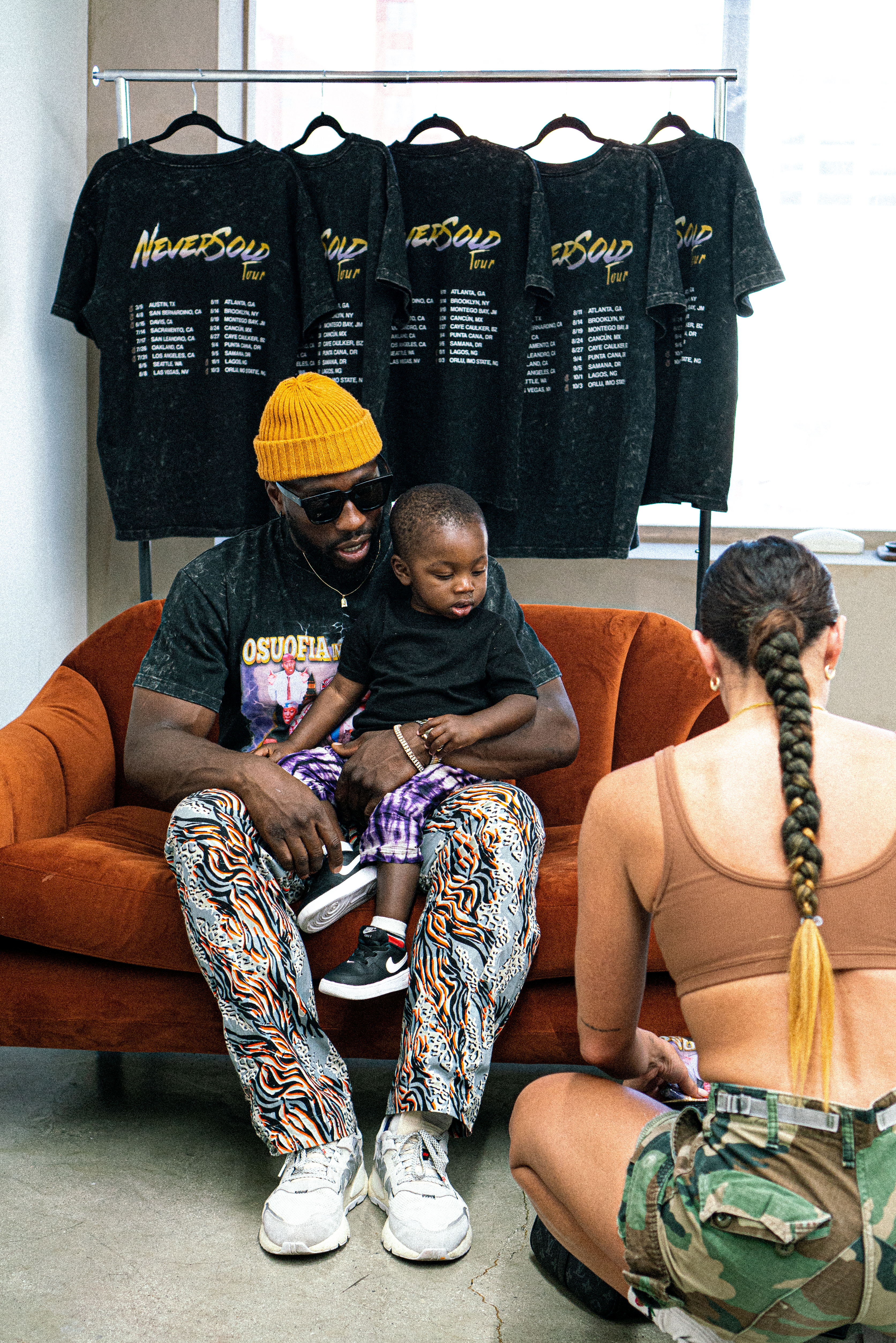Man in NeverSold Tour merch seated on orange couch with child and woman in studio