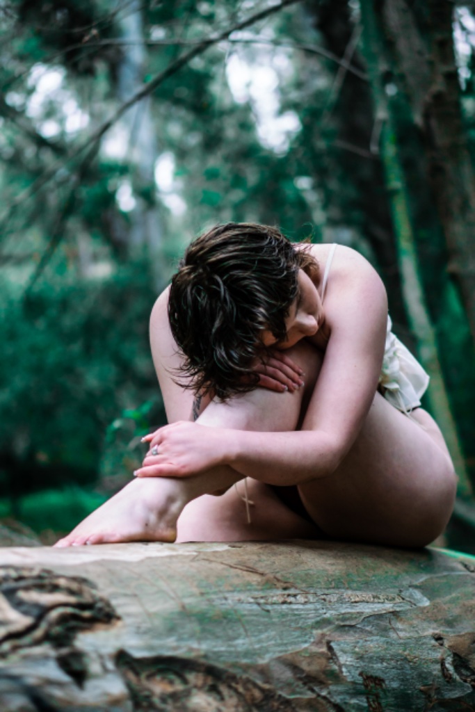 Woman in profile leaning forward on tree log, head bowed on knees in forest