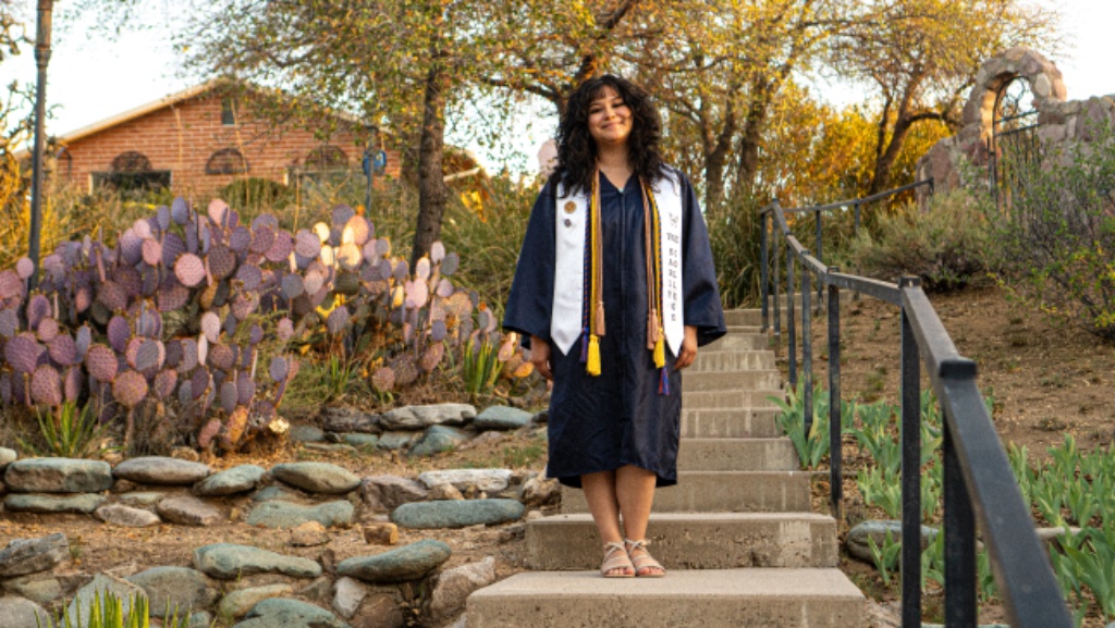 Graduate standing on outdoor concrete stairs with purple cacti and stone architecture