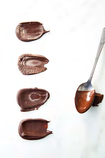 Four tempered chocolate samples smeared side by side on a pale surface beside a spoon, showing differences in set and sheen