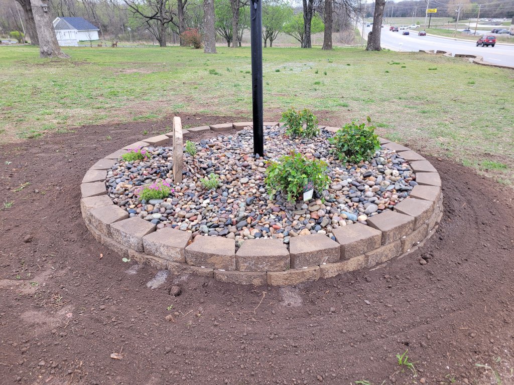 Circular hardscape bed with river rock and retaining wall blocks around flagpole