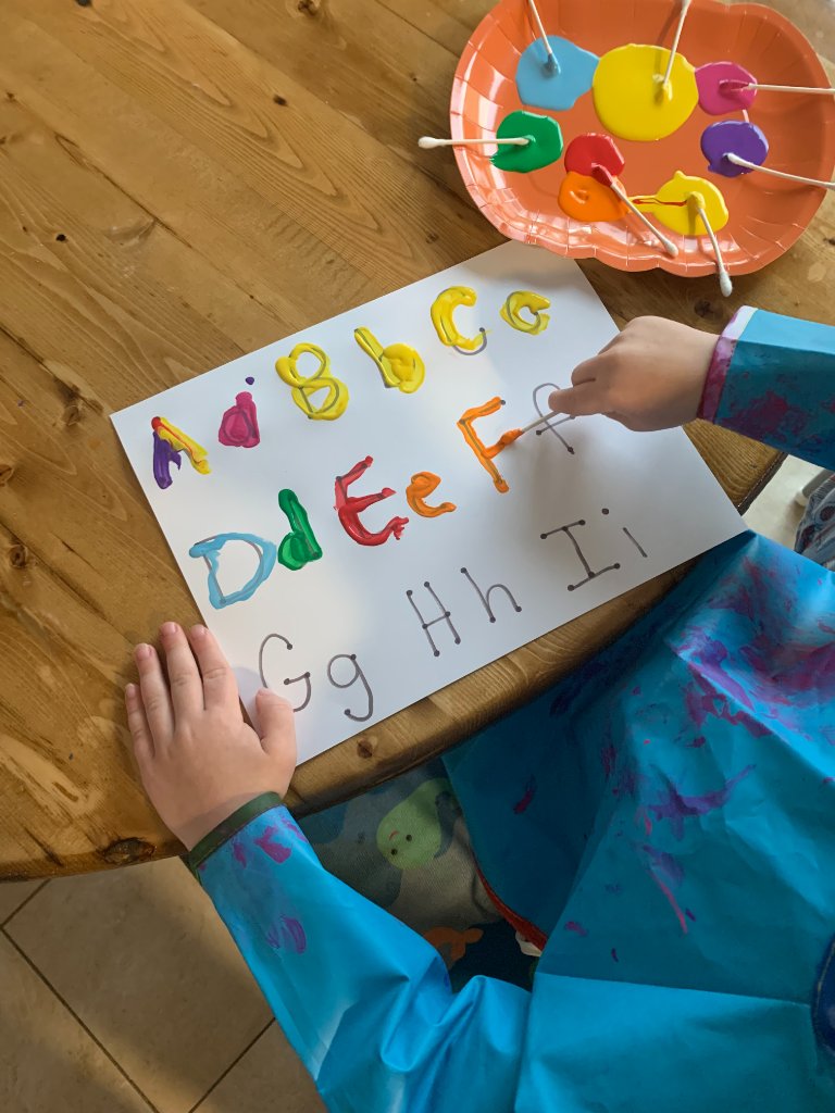 Child in a blue art smock painting alphabet letters with cotton swabs at a wooden table
