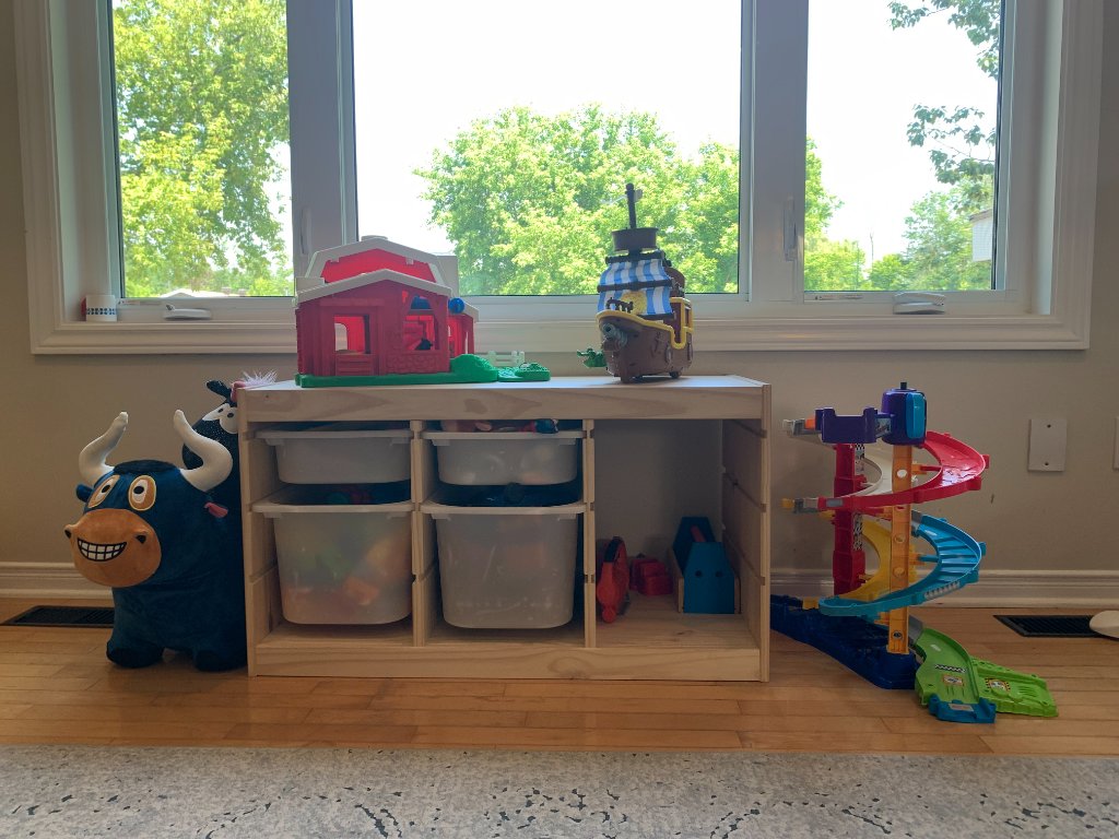 Wooden toy storage unit with bins, toy barn, and spiral car ramp near a bright window