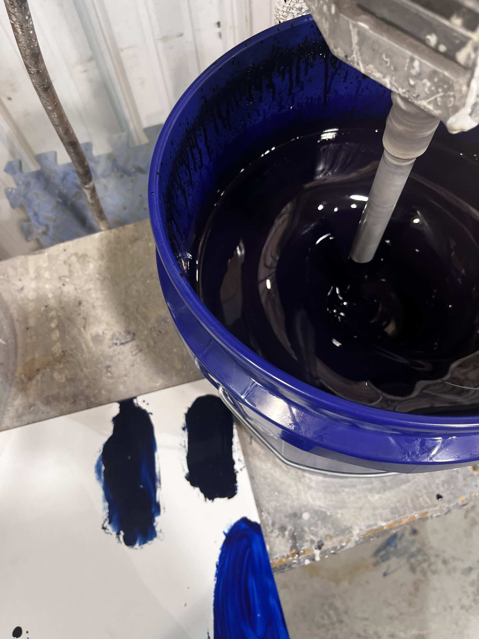 A blue bucket of dark pigment being mixed by a mechanical agitator, with test swatches nearby.