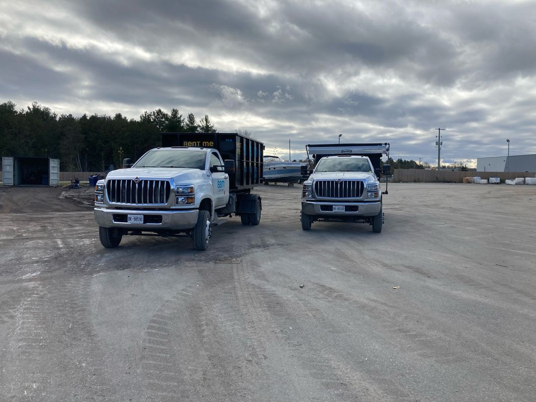 Two white BAM dump trucks parked on gravel under a cloudy sky