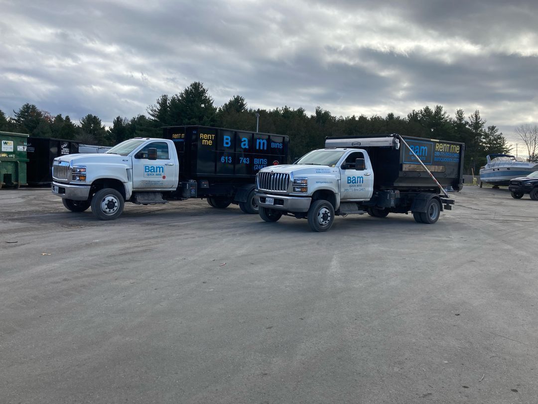 Two white BAM dump trucks parked on a gravel lot