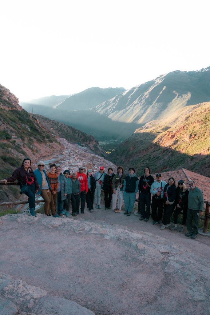 Vivara retreat group posing together along a wooden fence overlooking the Maras Salt Mines in the Sacred Valley of Peru, with terraced salt pools and canyon mountains at golden hour