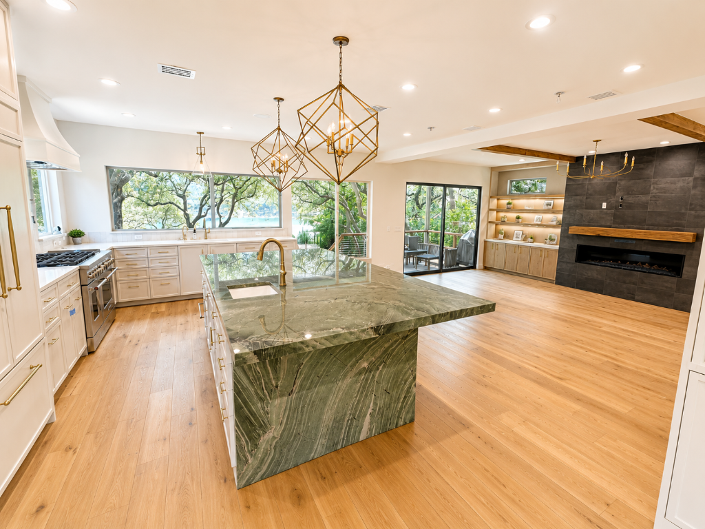 Open-concept kitchen with green-veined marble waterfall island, white shaker cabinetry, gold cage chandeliers, and a charcoal tile fireplace feature wall overlooking Lake Travis