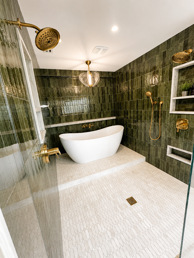 Modern wet room with freestanding soaking tub, frameless glass shower, mossy green vertical subway tile, pebble stone floor, and brushed brass fixtures