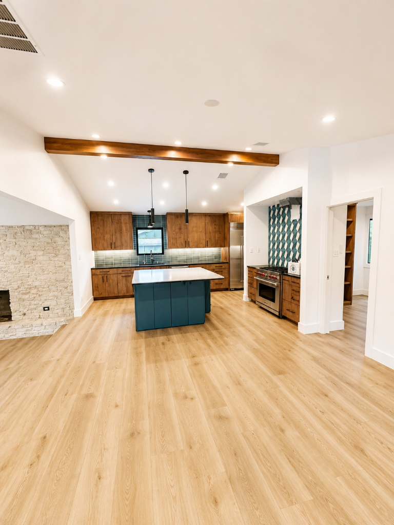 Open-concept kitchen with stone fireplace, deep teal island, medium wood cabinetry, patterned tile backsplash, and exposed wood beam on a vaulted ceiling
