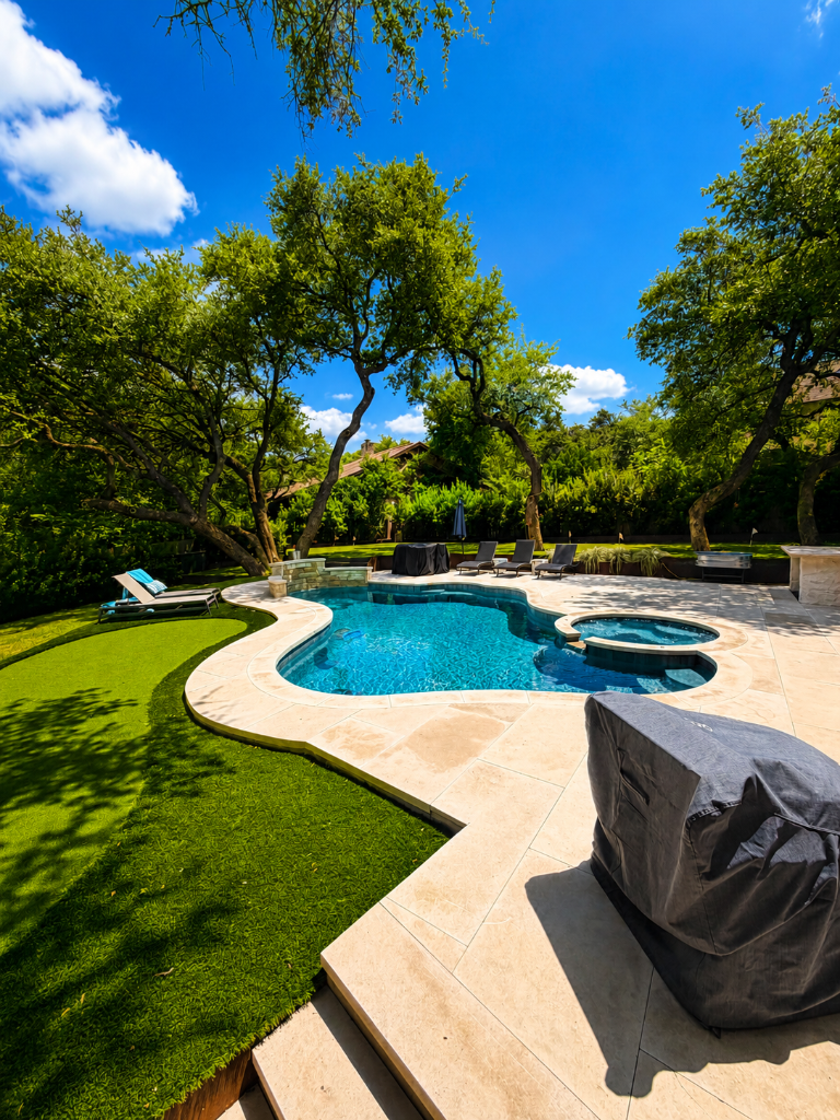 Curved pool with travertine decking, artificial turf, and lounge area surrounded by mature trees
